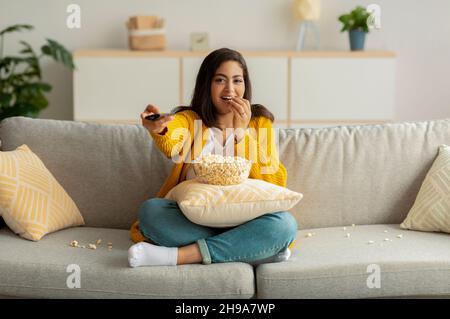 Junge arabische Frau, die mit Fernbedienung auf der Couch sitzt, sich einen Film anschaut und Popcorn isst und Freizeit genießt Stockfoto