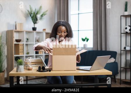 Aufgeregte afroamerikanische Frau, die am Schreibtisch saß und den Paketkasten öffnete. Weibliche Käuferin, die bestellte Waren vom Lieferservice überprüft. Konzept von Menschen und Einkäufen. Stockfoto