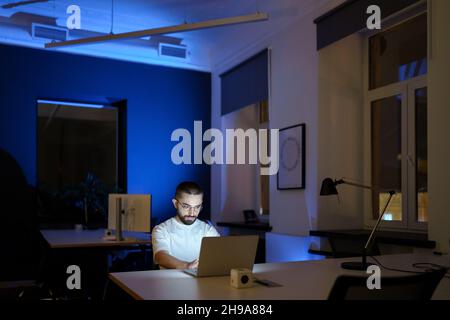 Konzentrierter Mann Web-Designer in Brillen Aufenthalt im Büro Überstunden Arbeit an Projekt in der dunklen Nacht Stockfoto