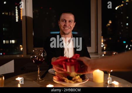 valentinstag bei Kerzenschein, weibliche Hand gibt ein Geschenk an einen Mann am Tisch sitzen, romantische Datum, Porträt eines Mannes in der Liebe in einem Anzug mit Wein Stockfoto