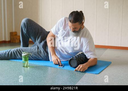 Kaukasischer bärtiger, langhaariger Mann mit Schaumstoffrolle und Shaker mit Wasser auf dem Yoga-Teppich und Tippen auf seinem Handy. Online-Schulungskonzept. Stockfoto