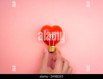 Hand halten Glühbirne Symbol Symbol auf roten Herzkugel auf rosa pastellfarbenen Hintergrund, minimalistischen Stil. Liebe, Fürsorge, Teilen, Geben, Wohlbefinden, Inspiration, A Stockfoto