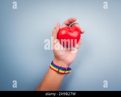 Hand mit kleinem Regenbogenfahnengürtel, der rote Herzkugel auf blauem Hintergrund hält und zeigt. Das LGBT-Konzept mit stolzen Farben und Regenbogenflaggenstreifen. Stockfoto