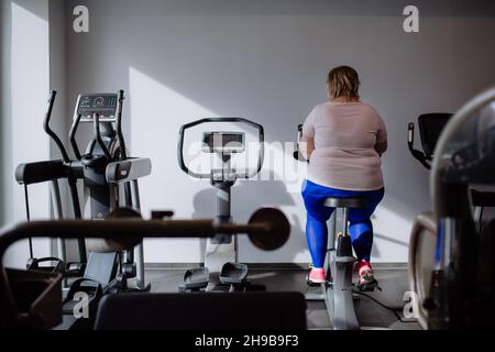 Rückansicht einer übergewichtigen Frau mit mittlerem Erwachsenen, die im Fitnessstudio auf einem stationären Fahrrad trainiert Stockfoto