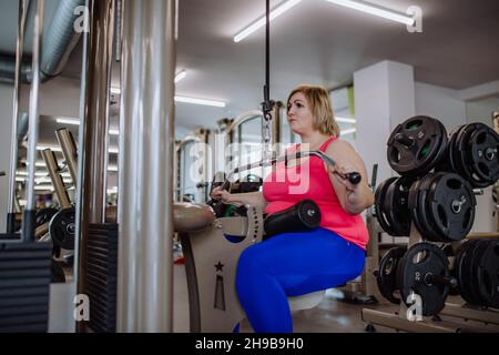 Mittlere Erwachsene plus große Frau, die im Fitnessstudio an einem Latenzausziehgerät trainieren kann Stockfoto