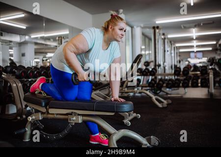Übergewichtige Frau mit mittleren Erwachsenen, die im Fitnessstudio mit Hanteln auf der Bank trainiert Stockfoto