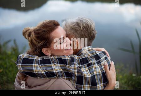 Rückansicht einer glücklichen älteren Mutter, die sich mit einer erwachsenen Tochter umarmt, wenn sie am See in der Natur sitzt Stockfoto