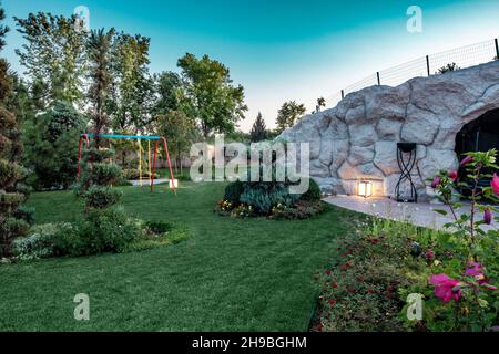 Sommerlandschaft von Parklandschaft mit Kinderschaukel auf grünem Rasen mit Blumenbeeten im Sommer Stockfoto