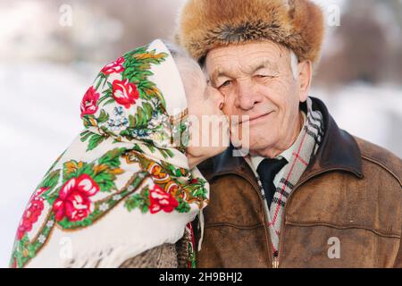 Porträt eines glücklichen älteren Paares. Ältere Frau küssen ihren Mann in gewichtigen. Altes Paar zu Fuß im Park im Winter Zeit. Frohe Familie. Goldene Hochzeit Stockfoto