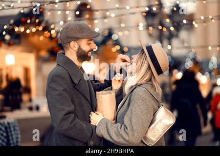 Romantisches Weihnachtspaar, das am Weihnachtstag auf der Straße Popcorn füttert. Ein junger glücklicher Mann füttert seine liebende Freundin mit Popcorn, während Stan Stockfoto