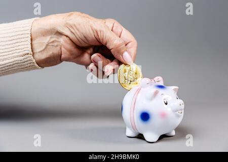 Faltige Hand einer älteren Frau legt eine Bitcoin in ein Sparschwein. Grauer Hintergrund. Das Konzept von Einsparungen und Kryptowährung. Stockfoto