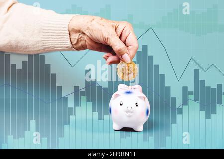 Faltige Weibchen legen eine Bitcoin ein Sparschwein. Business Chart im Hintergrund. Das Konzept von Investitionen und Finanzen. Stockfoto