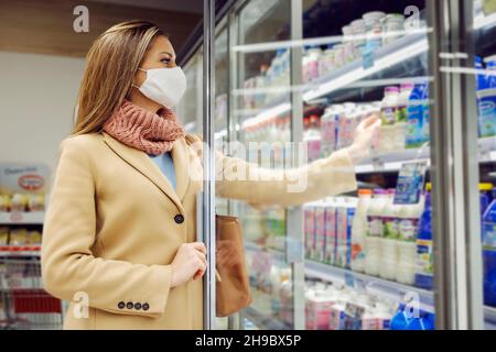 Eine Frau mit Gesichtsmaske nimmt frische Milch aus dem Kühlschrank im Laden. Frau, die Lebensmittel einkauft. Stockfoto