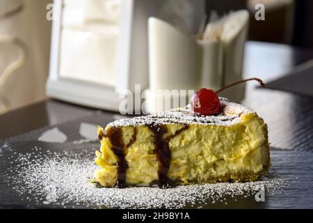 Ein Stück Käsekuchen mit Kirschen auf schwarzem Steinbrett auf dem Tisch im Café. Nahaufnahme. Selektiver Fokus. Stockfoto