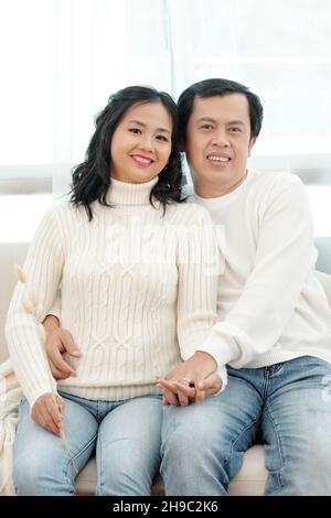 Glücklicher Mann und Frau in warmen Pullovern und Jeans sitzen zu Hause auf der Couch und lächeln vor der Kamera Stockfoto