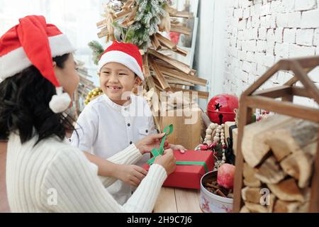 Lächelnder präjugendlicher Junge, der Mutter hilft, das Weihnachtsgeschenk zu verpacken und zu schmücken Stockfoto