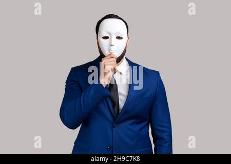 Anonymer unbekannter Geschäftsmann, der ihr Gesicht mit einer weißen Maske bedeckt, ihre wahre Persönlichkeit, Anonymität und ihren offiziellen Anzug versteckt. Innenaufnahme des Studios isoliert auf grauem Hintergrund. Stockfoto