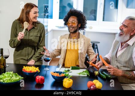 Glückliche Erwachsene Freunde kochen und bereiten veganes Abendessen zu, Menschen haben Spaß beim Singen, Lachen und Vorgeben, einige Musikinstrumente zu spielen Stockfoto