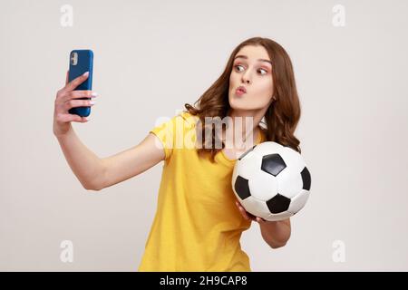 Junge attraktive Teenager Mädchen in gelbem Casual-Stil T-Shirt machen Selfie, mit Videoanruf oder Übertragung Livestream mit Ball in der Hand. Innenaufnahme des Studios isoliert auf grauem Hintergrund. Stockfoto