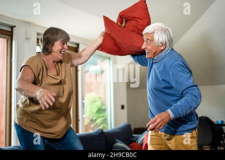 Spielerisches und sorgloses Seniorenpaar, das Kissen macht, kämpft mitten im Wohnzimmer, Freizeit-, Lifestyle- und Glückskonzept im Silberzeitalter Stockfoto