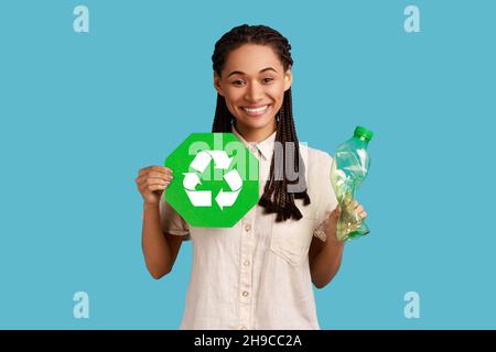 Glücklich zufriedene Frau mit schwarzen Dreadlocks halten in der Hand Recycling-Schild und Plastikflasche, Mülltrennung, trägt weißes Hemd. Innenaufnahme des Studios isoliert auf blauem Hintergrund. Stockfoto