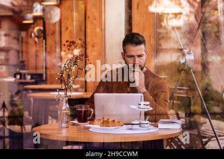 Schöner Mann in legerer Kleidung, während er im Café sitzt und am Laptop arbeitet. Stockfoto