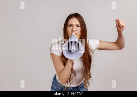 Porträt einer Frau, die das Megaphon in der Nähe des Mundes hält, laut spricht, schreit, mit erhobenem Arm Ansage macht, protestiert, weißes T-Shirt trägt. Innenaufnahme des Studios isoliert auf grauem Hintergrund. Stockfoto