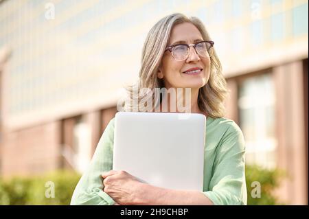 Nachdenkliche Frau in Brille umarmt Laptop auf der Straße Stockfoto