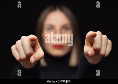 Nahaufnahme eines Frauenporträts, das die Finger auf die Kamera zeigt, mit Brille, roten Lippen, auf schwarzem Hintergrund und fokussiertem Gesicht an den Fingern. Du bist der Gewinner, ich werde dich finden. Wir suchen Sie Stockfoto