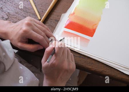 Der Künstler zeichnet mit einem Kunstpinsel bunte Farbverläufe in Aquarellfarbe auf eine weiße Leinwand. Blick hinter die Schulter. Zeichnung mit Aquarellen in einem Stockfoto