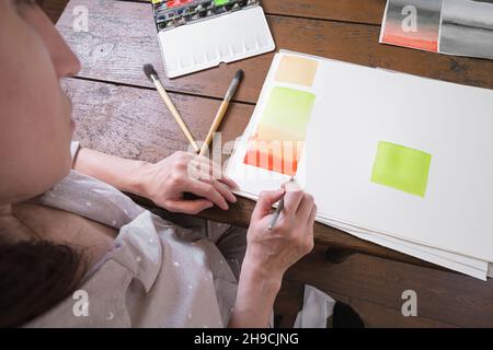 Der Künstler zeichnet mit einem Kunstpinsel bunte Farbverläufe in Aquarellfarbe auf eine weiße Leinwand. Blick hinter die Schulter. Zeichnung mit Aquarellen in einem Stockfoto