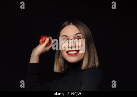 Aufgeregt Frau trägt Brille mit schwarzen Rahmen, mit roten Lippen, Blick auf die Kamera will einen roten Apfel über schwarzen Hintergrund werfen. Stockfoto