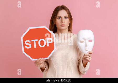 Porträt einer ernsthaften blonden Frau mit rotem Stoppschild und weißer Maske, die mit strengem Ausdruck die Kamera anschaut und einen weißen Pullover trägt. Innenaufnahme des Studios isoliert auf rosa Hintergrund. Stockfoto