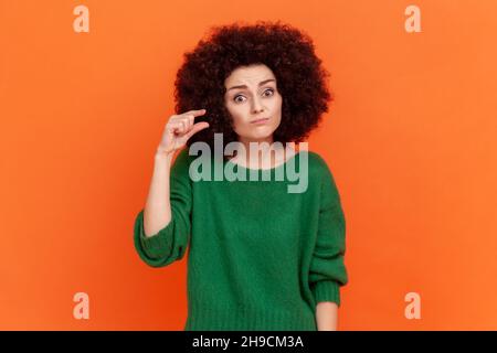 Porträt einer Frau mit Afro-Frisur, die einen grünen Pullover im lässigen Stil trägt und eine kleine Geste mit den Fingern zeigt, wobei sie enttäuscht auf die Kamera blickt. Innenaufnahme des Studios isoliert auf orangefarbenem Hintergrund Stockfoto