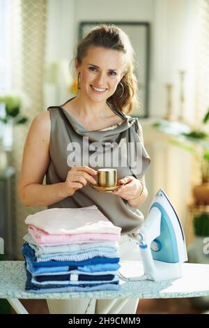 Fröhliche moderne Frau in Seidenbluse und beigen Hosen mit Dampfbügeleisen, Bügelbrett und Stapel von gefalteten gebügelten Kleidung trinken Tasse Espresso nach dem Hous Stockfoto