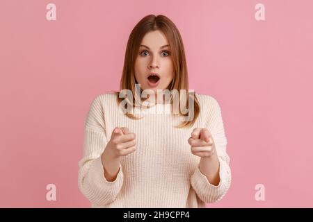 Porträt einer blonden Frau, die mit dem Finger auf die Kamera zeigt, schockiert mit offenem Mund, kaum zu glauben, mit weißem Pullover. Innenaufnahme des Studios isoliert auf rosa Hintergrund. Stockfoto