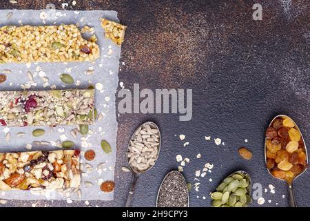 Verschiedene Müsliriegel mit Nüssen, Samen, Hafer, Beeren, trockenen Früchten und Löffeln mit Zutaten auf dunkelbraunem Hintergrund. Protein mues Stockfoto