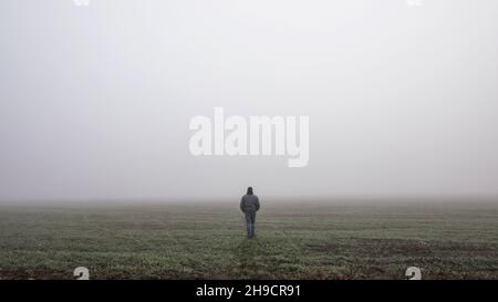 Ein einziger Mann geht in einer dramatischen mystischen Szene auf die neblige Straße. Kerl, der in einer nebligen Herbstlandschaft spazierengeht Stockfoto