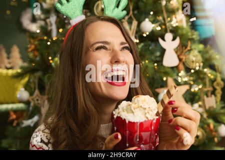 Weihnachtszeit. Glückliche elegante 40 Jahre alte Frau mit festlichem heißen Schokolade Cocktail in der Nähe von Weihnachtsbaum im modernen Haus. Stockfoto