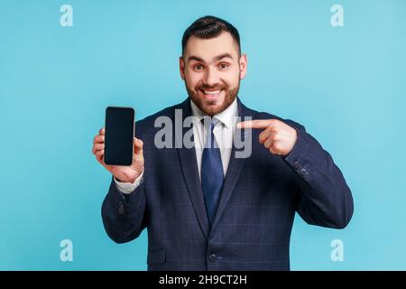 Fröhlicher, fröhlicher, perlter Mann in dunklem Anzug, der mit dem Finger auf das Smartphone zeigt und mit einem leeren Bildschirm die Kamera mit einem toothy Lächeln anschaut, Freiraum für adv. Innenaufnahme des Studios isoliert auf blauem Hintergrund. Stockfoto