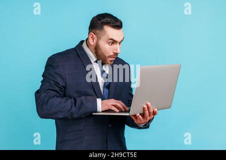 Schockiert Mann mit Bart tragen offiziellen Stil Anzug Blick auf Laptop-Display mit geöffnetem Mund und großen Augen, überrascht mit Betriebssystem-Fehler. Innenaufnahme des Studios isoliert auf blauem Hintergrund. Stockfoto