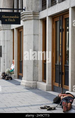 Ein Obdachloser schläft auf dem Boden vor dem Abercrombie & Fitch Store in München. An der Seite sitzt eine Dame mit ihrem Hund. Stockfoto