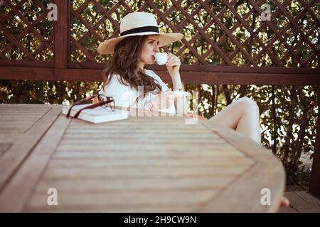Entspannte, stylische Hündin im weißen Hemd mit einer Tasse Kaffee, Buch, Hut und Brille, die auf der Terrasse des Gästehaushotels am Tisch sitzt. Stockfoto