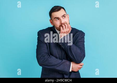 Ängstlich nervös Geschäftsmann mit Bart im offiziellen Stil Anzug beißen Nägel auf die Finger Blick auf die Kamera mit erschreckendem Ausdruck, besorgt über die Arbeit. Innenaufnahme des Studios isoliert auf blauem Hintergrund. Stockfoto