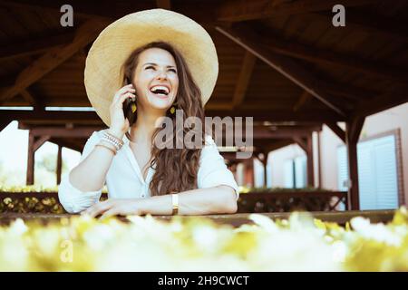 Glückliche trendige Frau mittleren Alters in weißem Hemd mit Hut mit einem Smartphone auf dem Bauernhof. Stockfoto