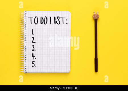 Offenes, spiralförmiges Notizbuch mit To-do-Liste und braunem Stift mit Hasenkopf auf hellgelbem Hintergrund, Draufsicht, flach liegend Stockfoto