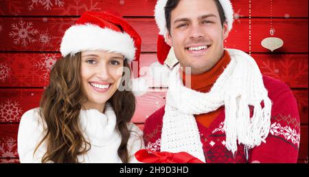 Porträt eines glücklichen jungen Paares, das weihnachtsmützen und warme Kleidung vor rotem Hintergrund trägt Stockfoto