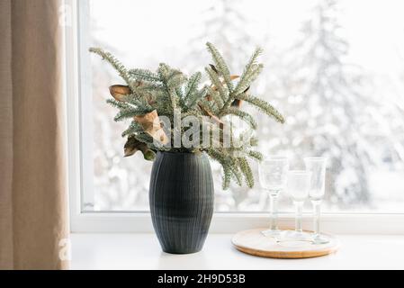 Weihnachtliche Komposition aus Christbaumzweigen in einer schwarzen Vase auf dem Fenster eines gemütlichen Hauses Stockfoto