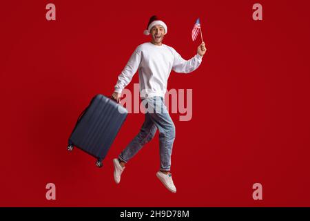 Aufgeregt Junger Mann In Santa Hat Springen Mit Amerikanischer Flagge Und Koffer Stockfoto