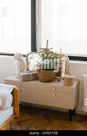 Ein Weihnachtsbaum und eine Geschenkbox in einem Korbkorb stehen auf einer hellen Kommode im Wohnzimmer oder Schlafzimmer im Inneren des Hauses Stockfoto